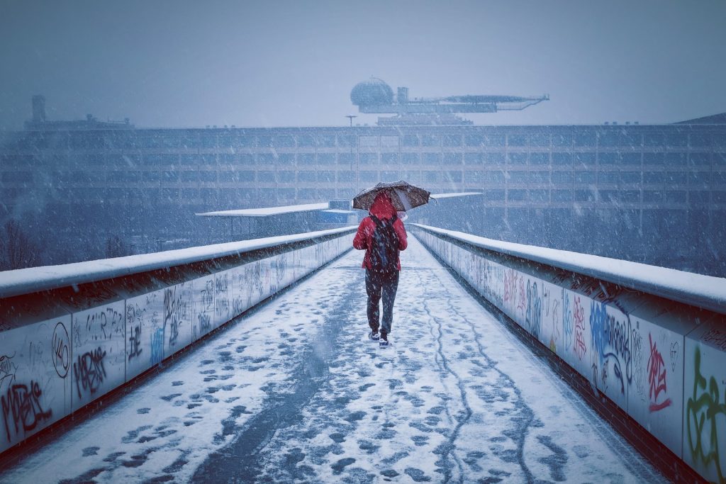 Person in a red hoodie walking down a snowy walkway, holding an umbrella against the falling snow.