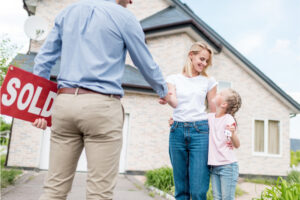 Real estate agent holding a "SOLD" sign and shaking hands with a woman and child in front of a house.